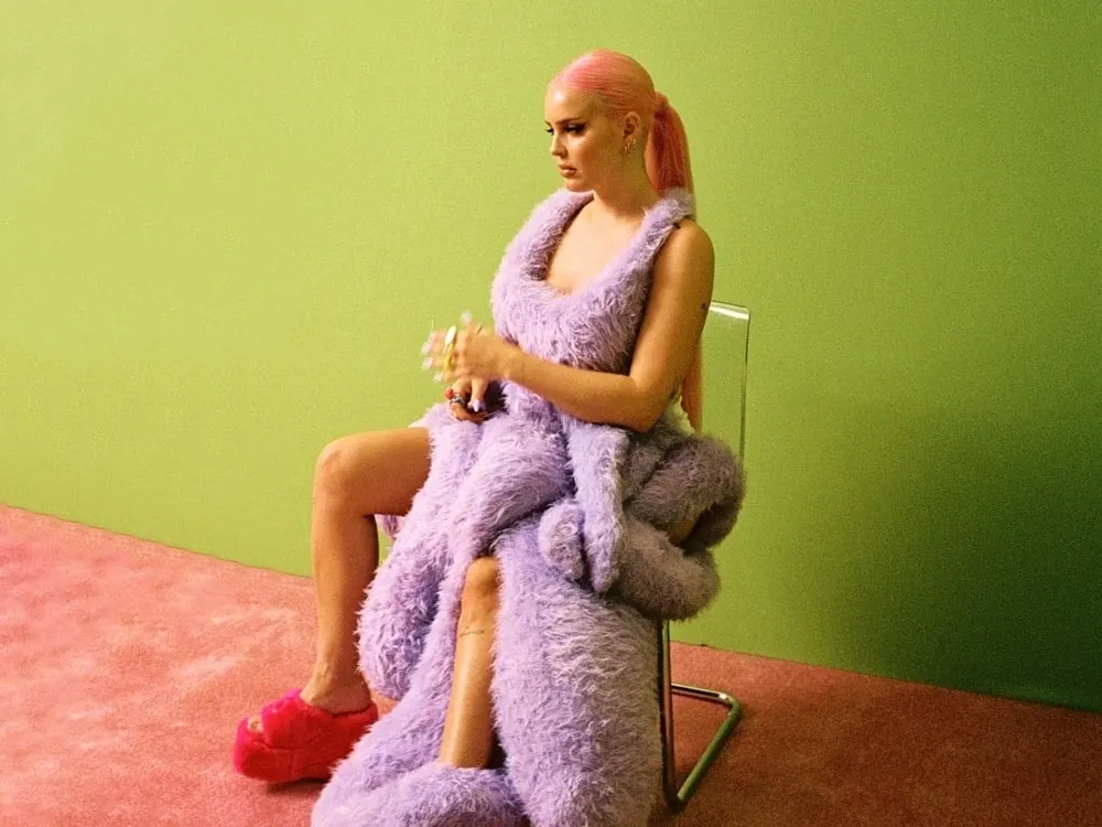 Anne-Marie in a lilac faux-fur wrap, seated against a lime-green wall