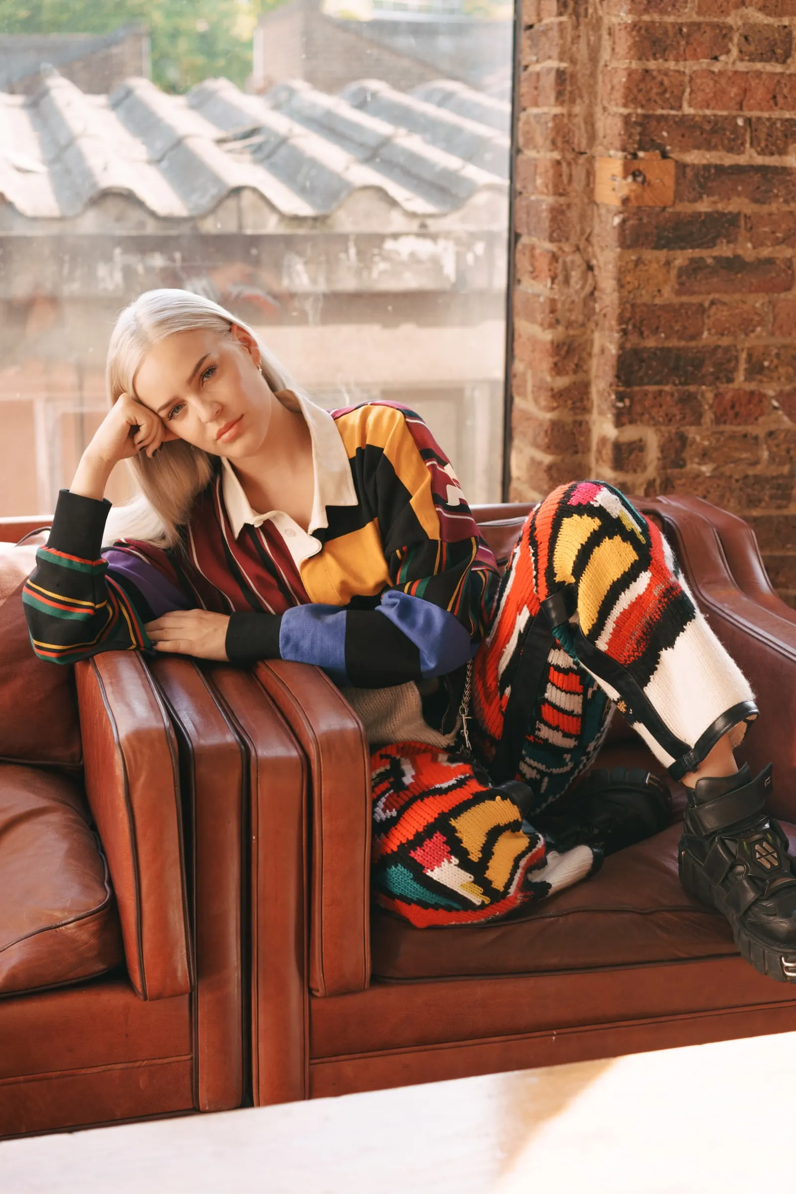 Anne-Marie lounging on a leather sofa in a bold knitted look, by a brick-walled window