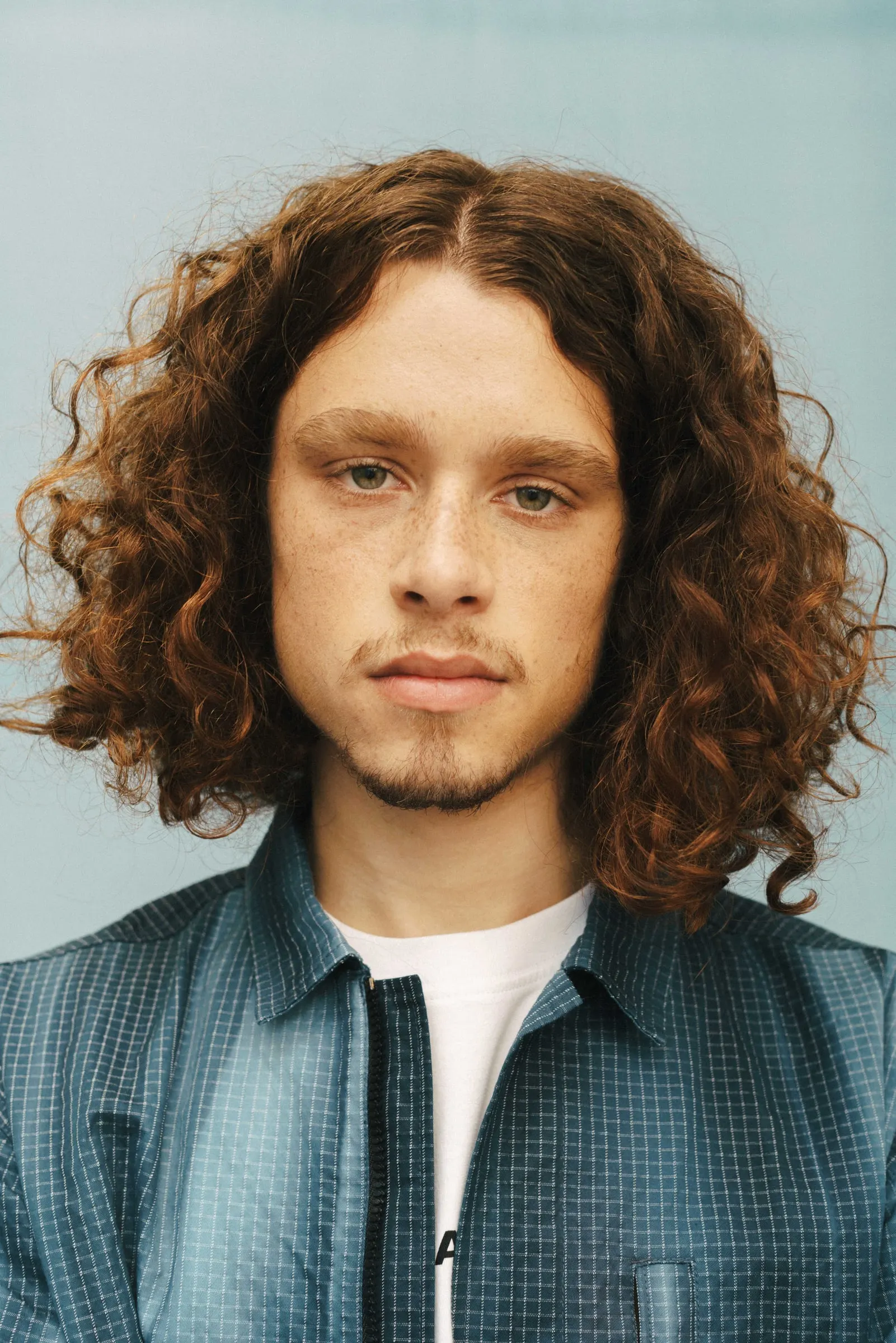 Close portrait of Izzy Cofie with long curly hair in a checked overshirt against a pale blue wall