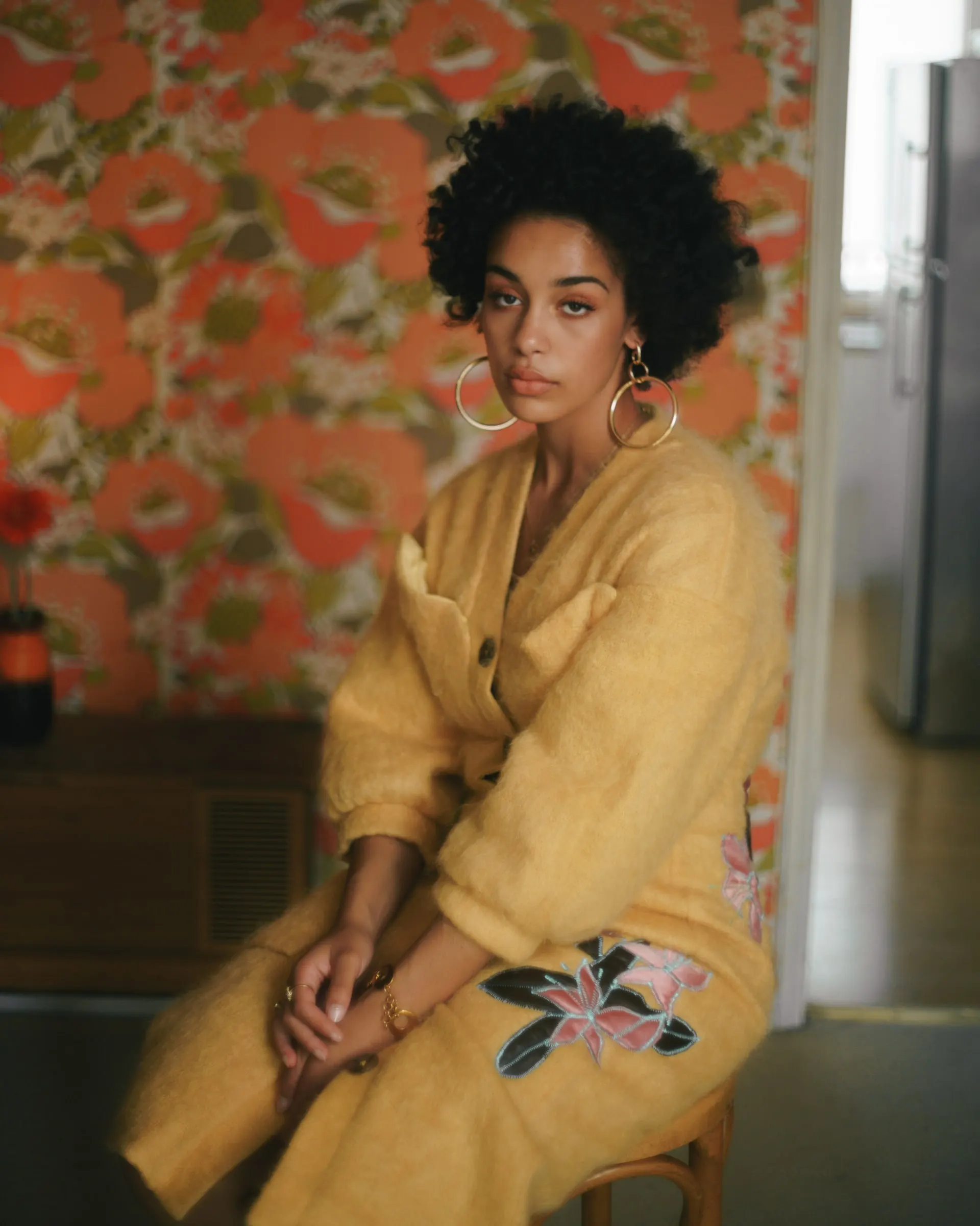 Jorja Smith in a yellow mohair cardigan, seated before orange floral wallpaper
