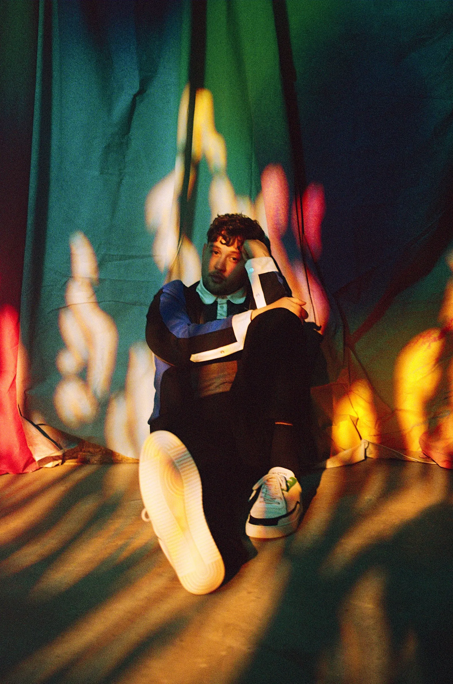 James Vickery resting his head on his hand, seated on the floor among projected patterns of coloured light