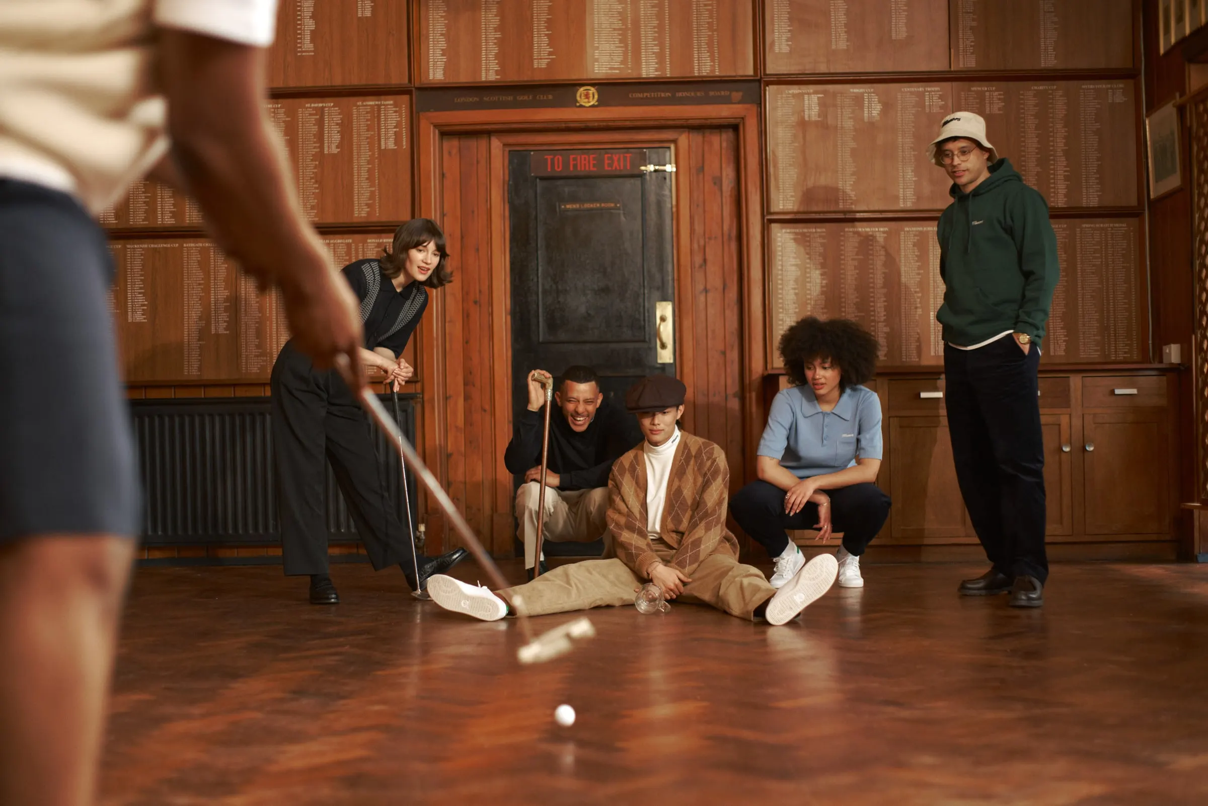 Friends in Manors gathered on a parquet floor, one putting a golf ball towards the camera