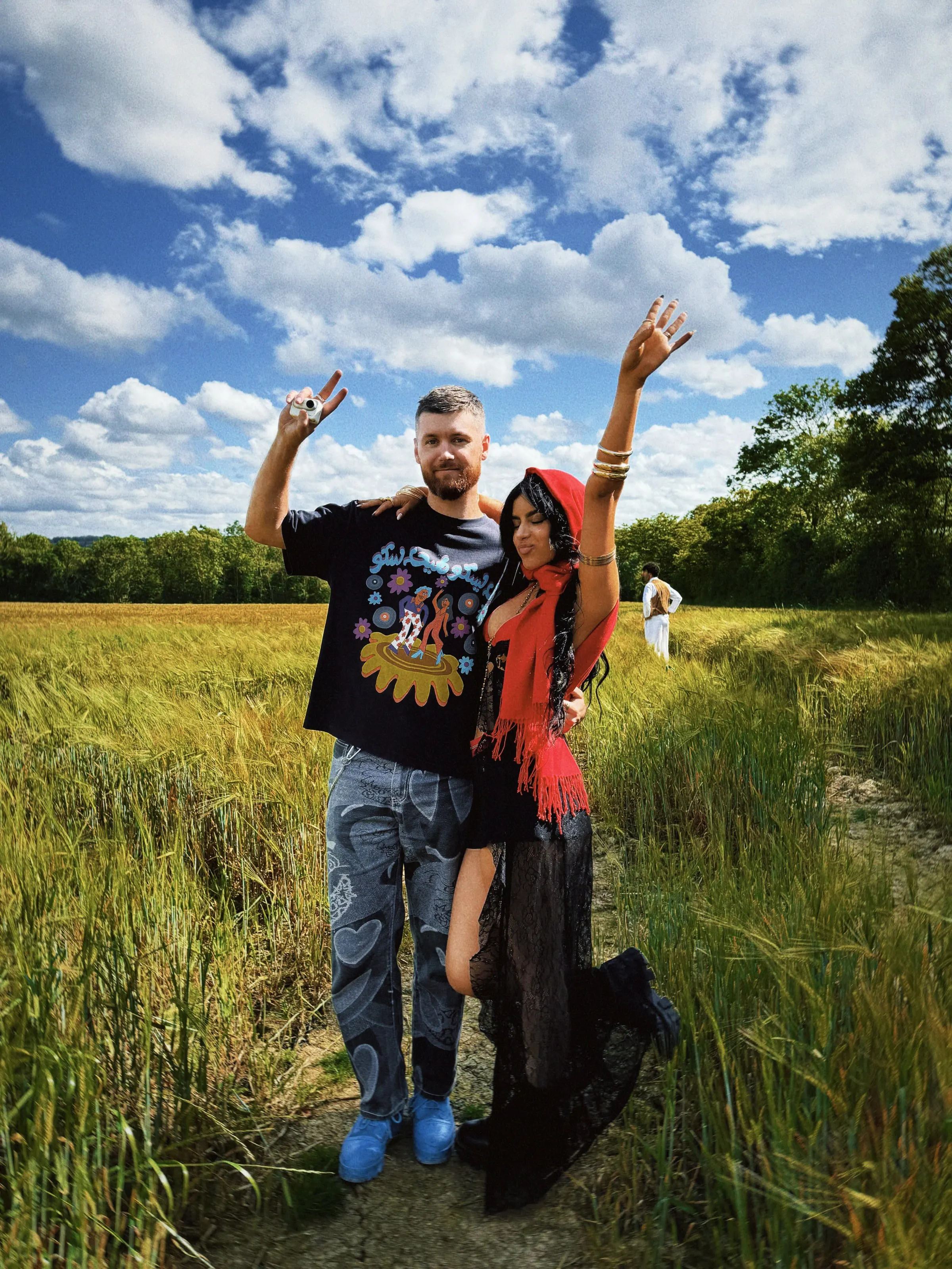 Will Beach and Maria Taktouk in the wheat field on the Always Go Back shoot: Maria in her red headscarf, both with an arm raised under a big blue sky