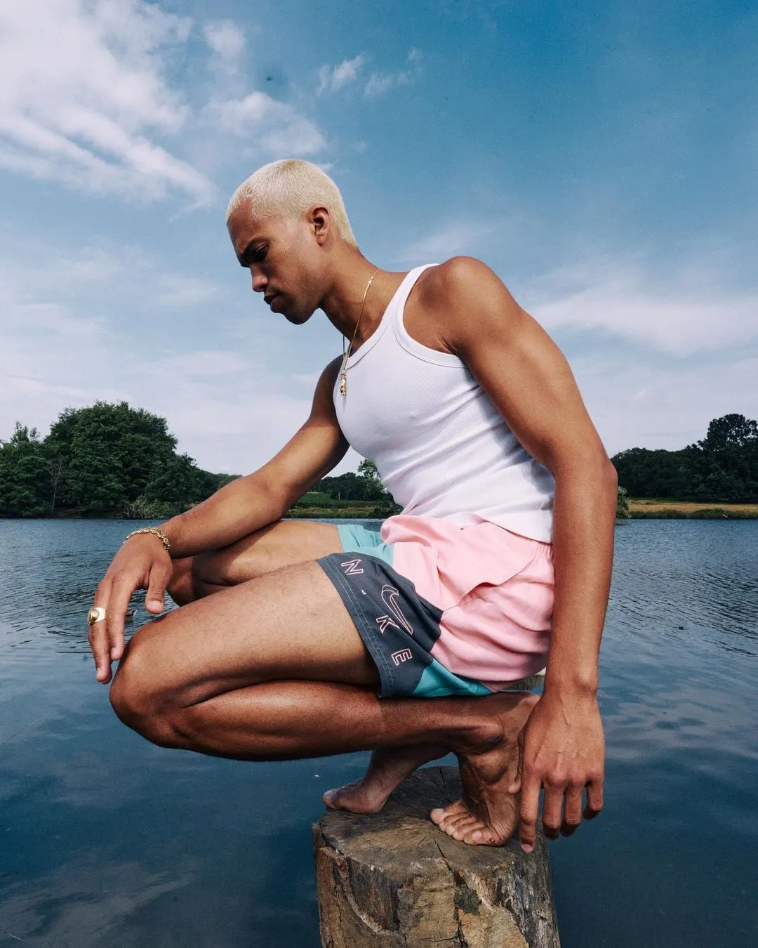 A man with bleached hair in a white vest and pink, teal and grey Nike swim shorts crouching barefoot on a tree stump above a lake