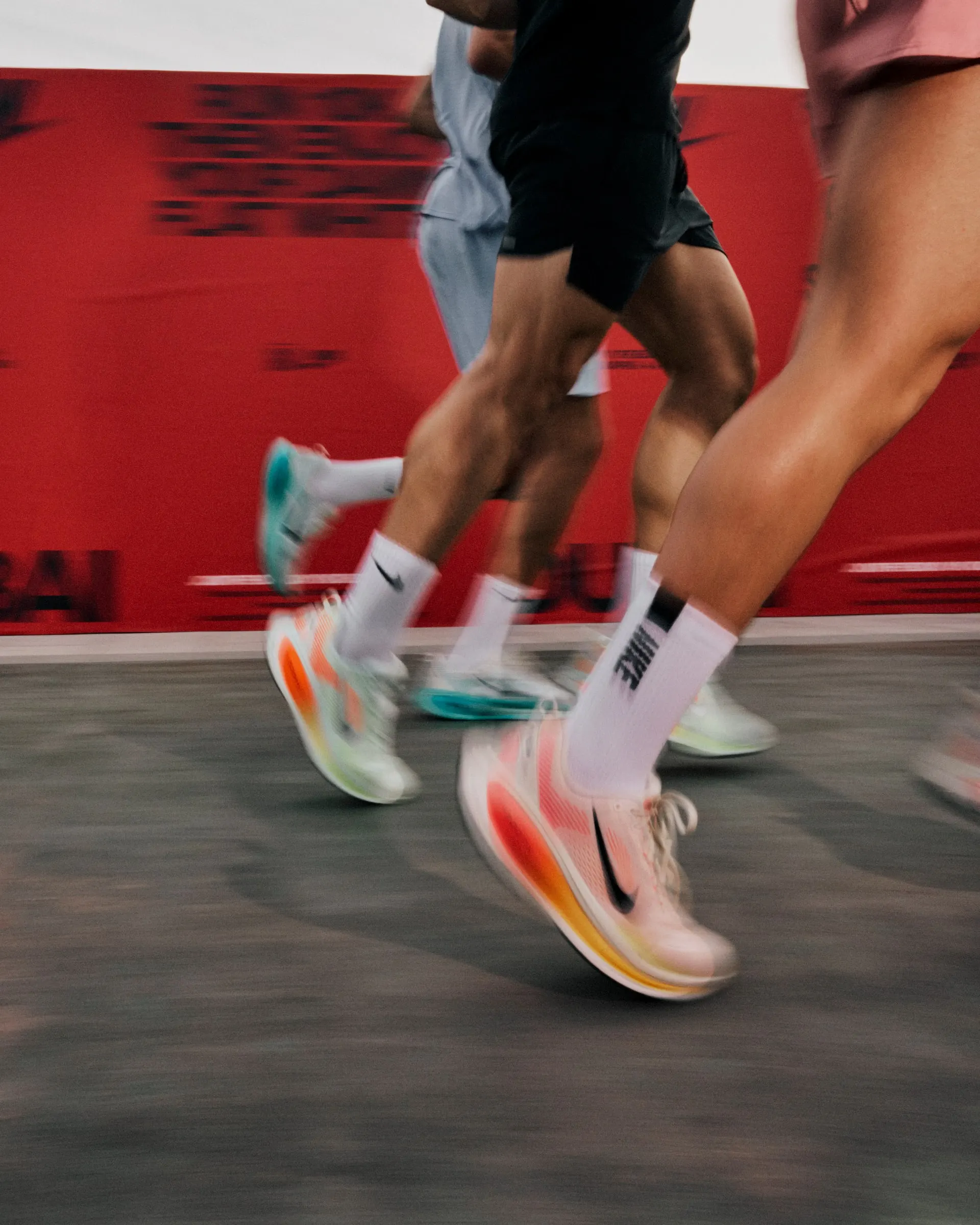 Runners in Vomero 18s blurred mid-stride against a red Nike wall