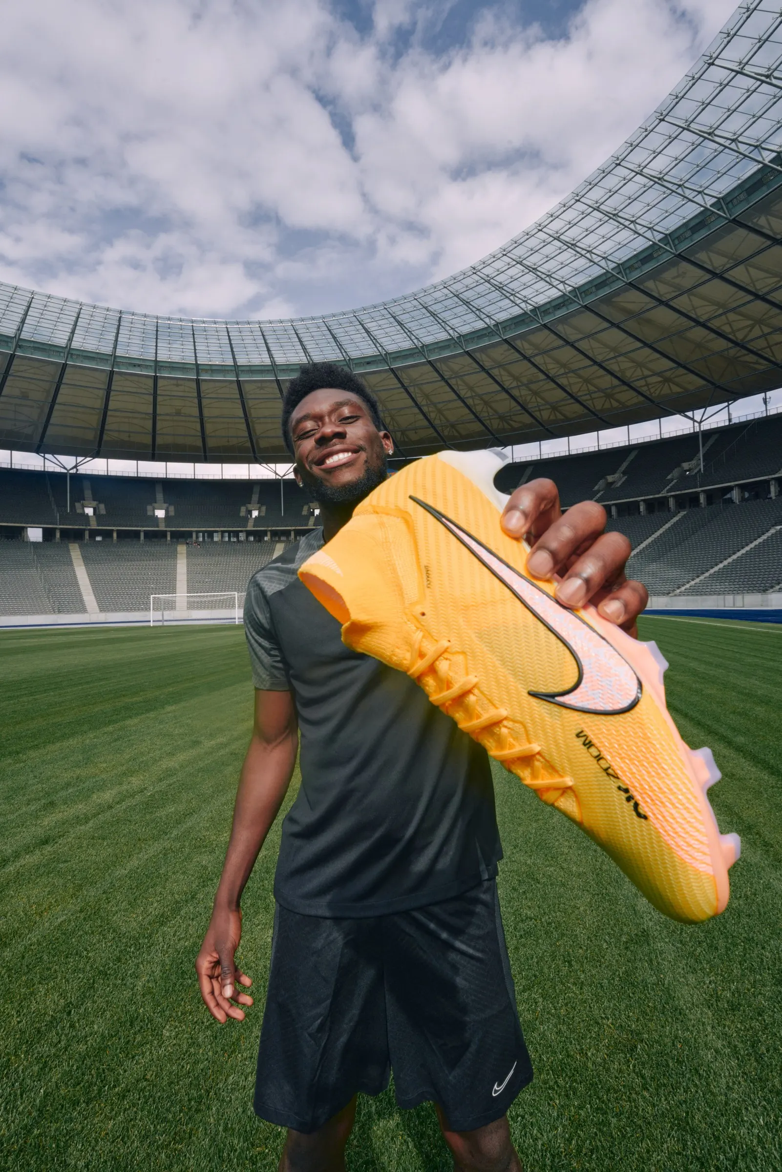 Alphonso Davies in the Olympiastadion, grinning as he holds a yellow Nike Mercurial out to the lens