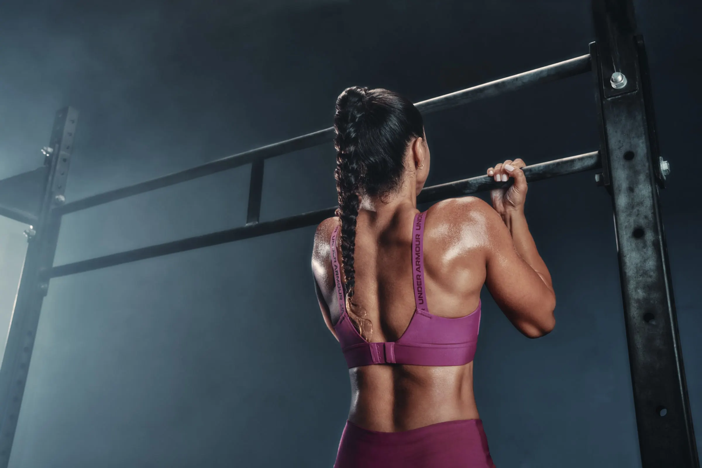A woman with a long braid gripping a pull-up bar in a smoky gym, shot from behind