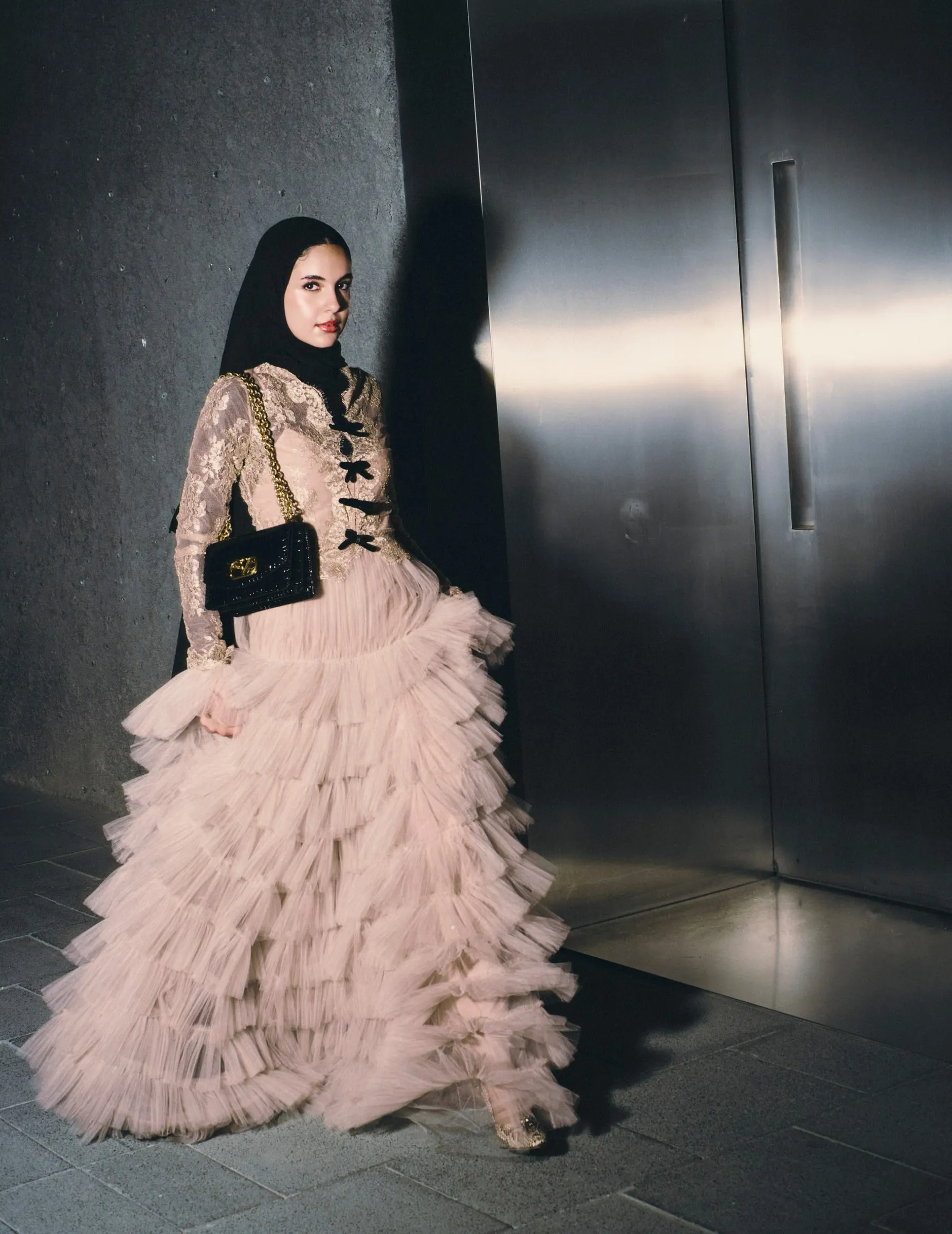 A model in a hijab and a tiered blush tulle gown against a brushed steel wall
